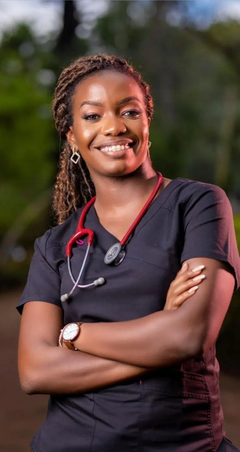 Dr Christine Nyabuto smiling, arms folded, in black scrubs with a red stethoscope around her neck, photographed outdoors against soft greenery.