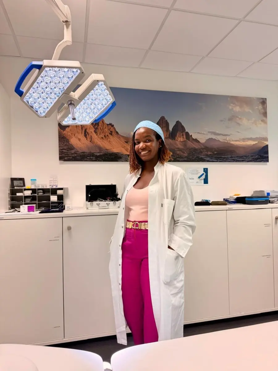 Dr Christine Nyabuto smiling in a white coat, pale pink top and magenta trousers, standing in a bright clinical room beneath an examination light.